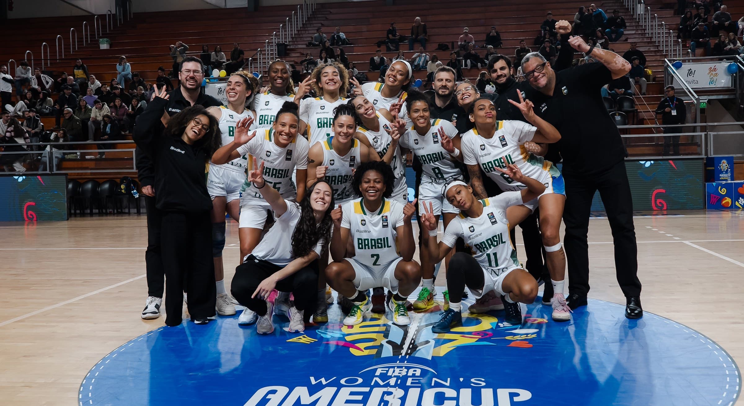 The Brazilian women’s national basketball team celebrating on court, with Joana Paiva among the staff.